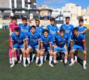 Group of 11 young soccer players in blue jerseys posing for a team photo on a sunny day, stadium stands in the background.