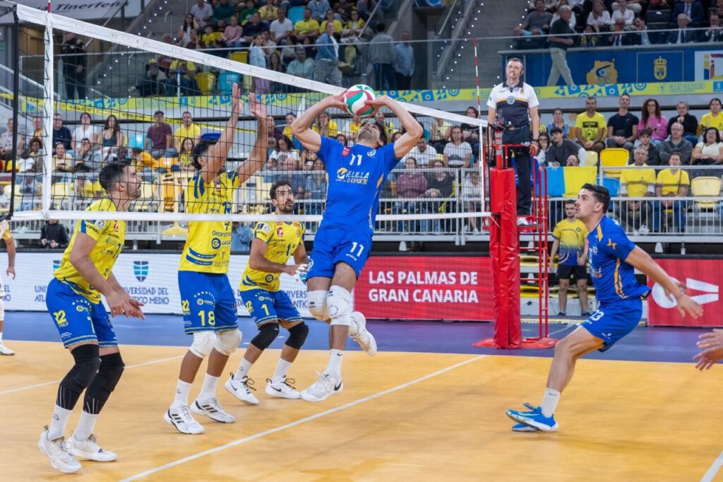 Blue volleyball player jumps to spike the ball over the net while yellow-clad blockers rise to defend, with spectators in the stands behind.