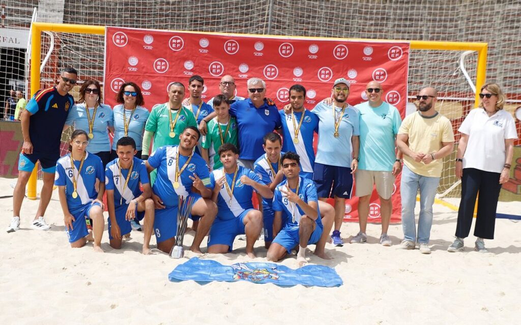 Group of beach soccer players and coaches posing on sand with medals and a trophy, in front of a red RF EF banner.