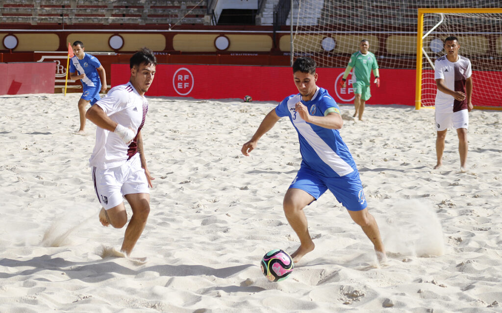 Beach soccer match with players in white and blue uniforms chasing a colorful ball on sandy pitch with a goal in the background