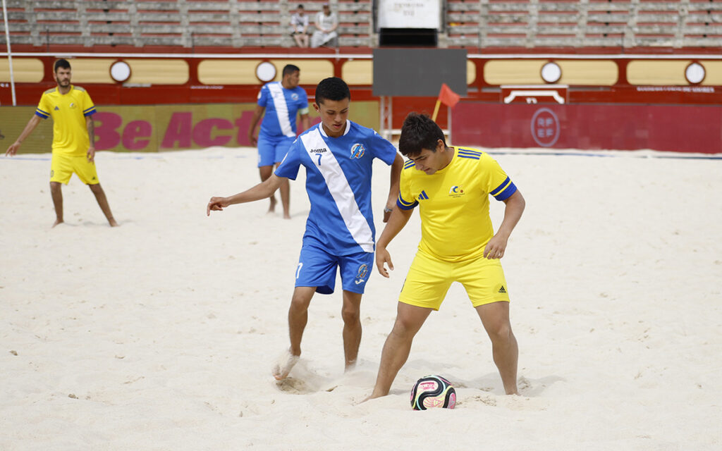 Two young players contest a ball on a sandy beach soccer field, one in blue and white and the other in yellow.