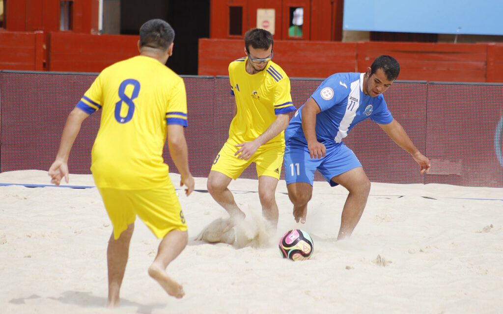 Three men play beach soccer: two in yellow uniforms and one in blue chasing a colorful soccer ball on sand with red barriers in the background.