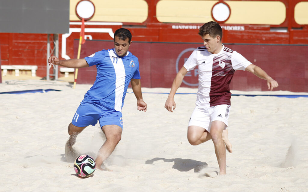 Two male beach soccer players battle for the ball on sand; a player in blue kicks while a rival in white and maroon pursues nearby.