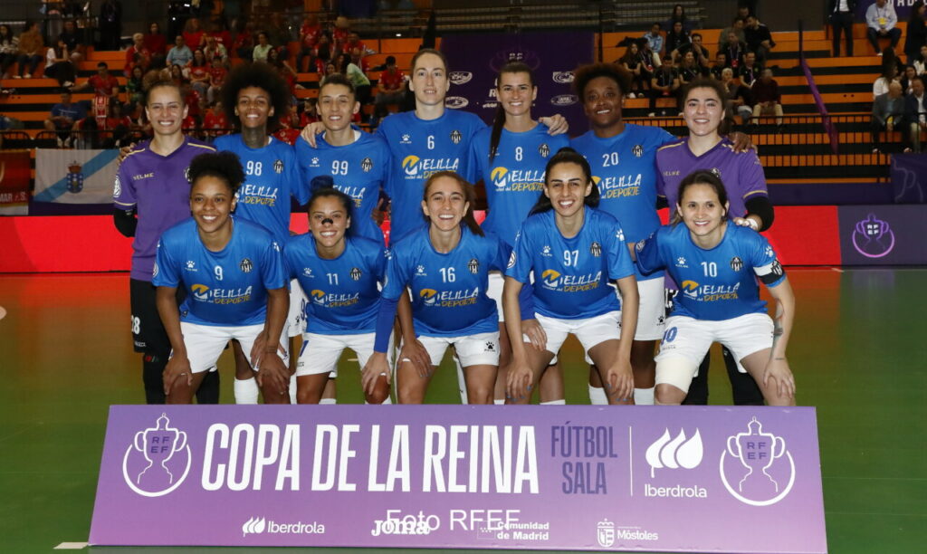 Group of female futsal players in blue jerseys posing for a team photo on an indoor court with a purple banner reading 'COPA DE LA REINA' in front.