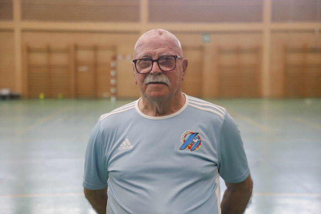 Older man with glasses and a mustache in a light blue Adidas sports jersey standing in a gymnasium, hands behind his back.