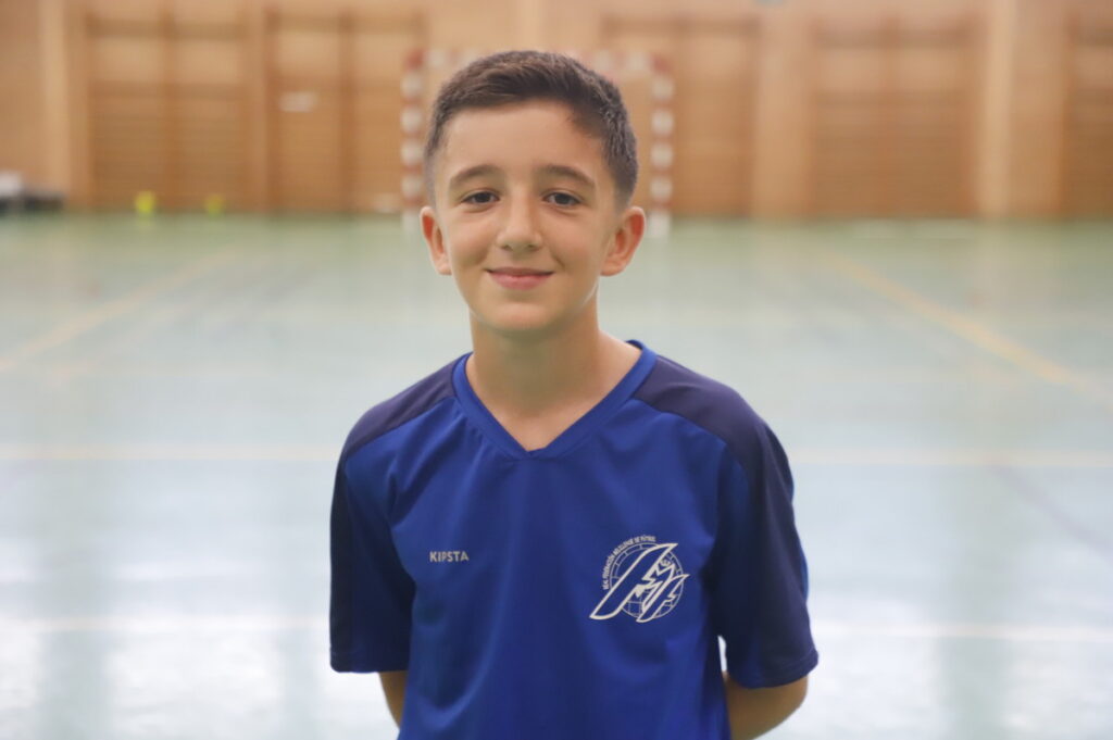 Smiling boy in a blue sports jersey standing in a gymnasium with a wooden wall in the background.