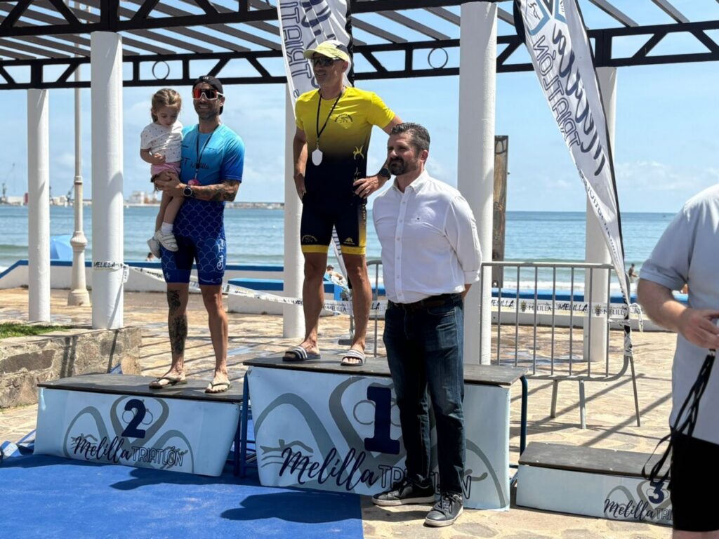 Three men stand on a seaside winners’ podium (1st place center in yellow/black, 2nd place left in blue holding a child, 3rd place right in white shirt) during a coastal triathlon award ceremony with banners and ocean in the background.