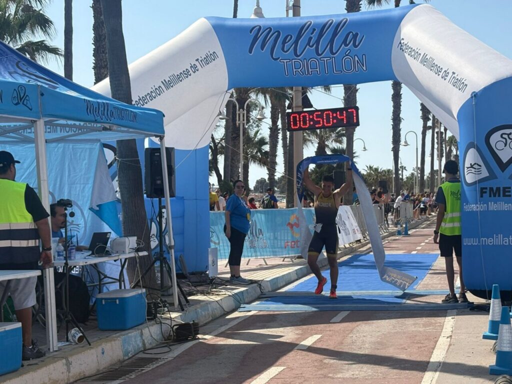 Athlete crosses the finish line under a blue and white arch that reads Melilla Triatlón, timer shows 00:50:47, on a palm-lined street.