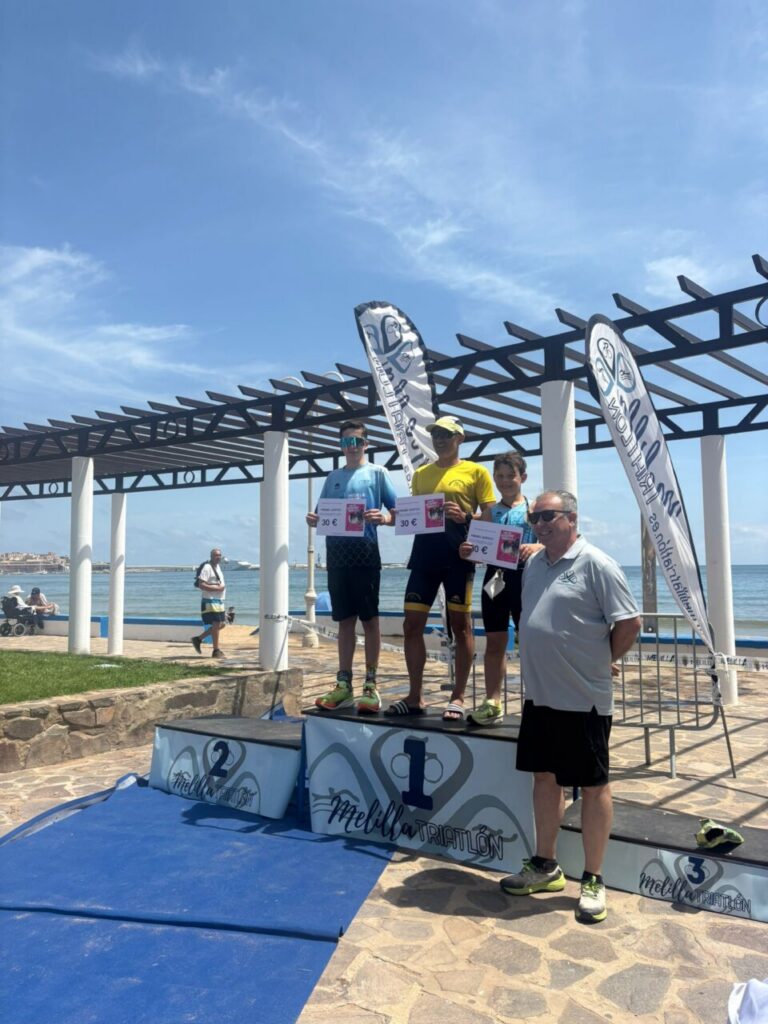 Three young athletes stand on a winners' podium by the sea, holding certificates as a coach watches nearby.