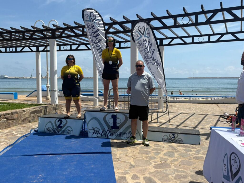 Two female athletes in yellow tops stand on the beachside winners' podiums with a man beside them, medals around their necks and the sea in the background.