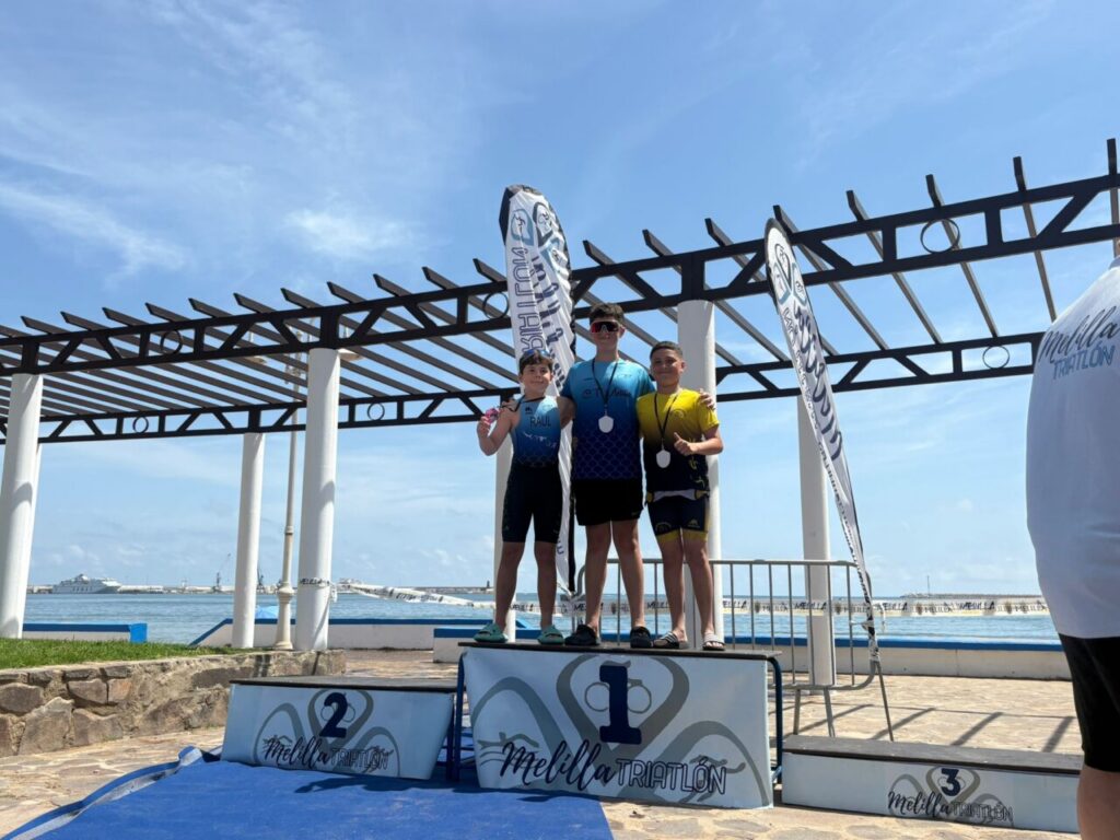 Three young athletes stand on a winners’ podium by the seaside, wearing medals and posing for a photo after a triathlon event at Melilla.