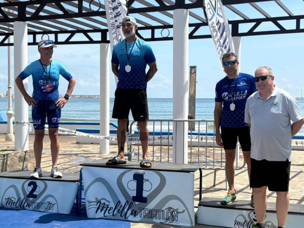 Winner stands on the central podium with a medal, flanked by second and third place on coastal podium with blue kits; ocean backdrop.