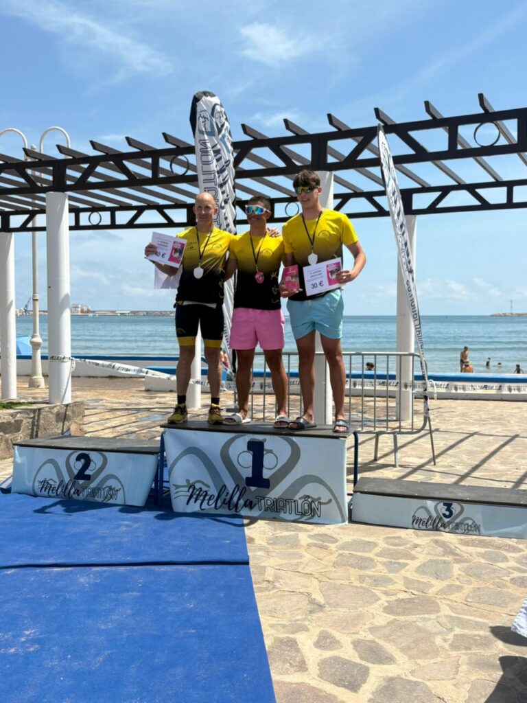 Three male podium finishers stand on a beach awards platform with medals and certificates, ocean behind them and a sunny sky. First, second, and third place markers are visible on the podiums labeled 1, 2, and 3. They wear yellow shirts and shorts, celebrating a triathlon event at the seaside.