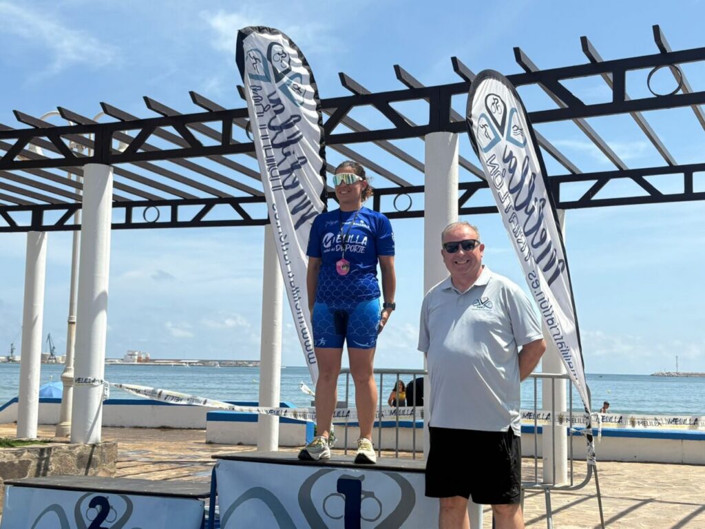 Female athlete on a beach podium wearing blue athletic gear and a medal, with a man in a polo shirt beside her under a steel framework. [sports event]
