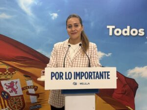 Woman at a lectern giving a speech, wearing a pink check blazer with the Spanish flag backdrop and a banner reading 'POR LO IMPORTANTE' in Melilla.