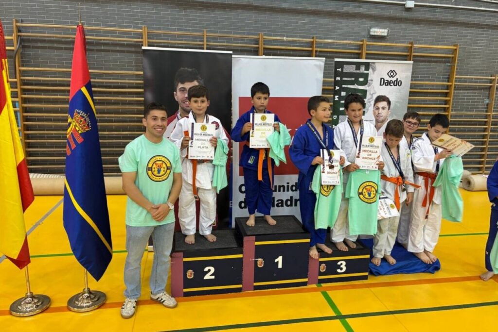 Group of young martial artists on a winners' podium, holding certificates and towels, with a smiling coach beside and flags in the gym background.