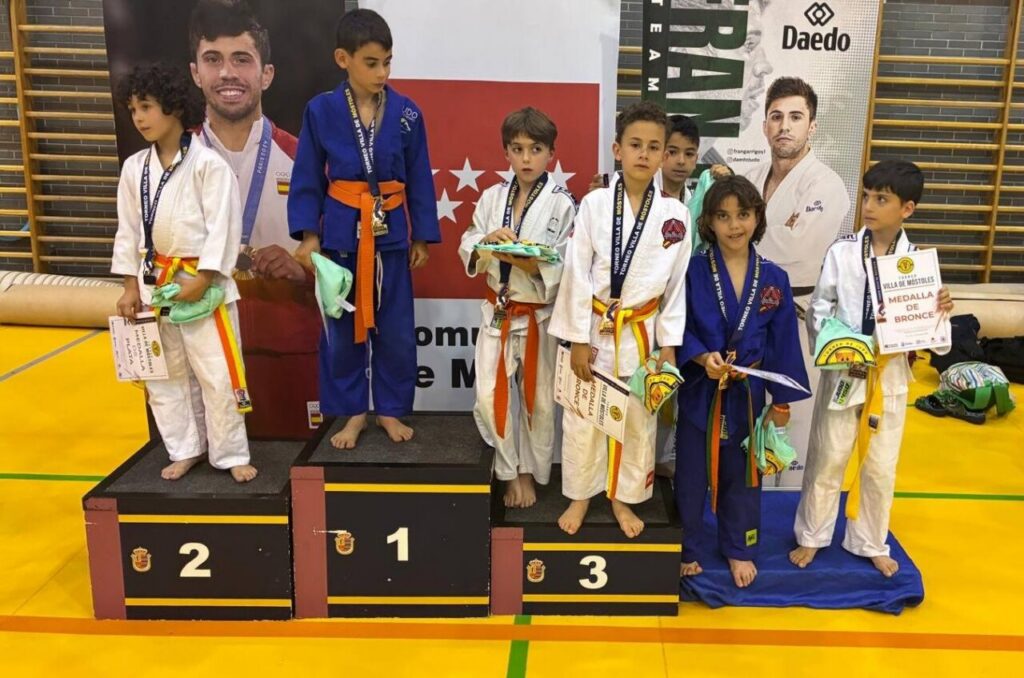 Young judo competitors on a winners' podium, holding medals and certificates with banners in the gym backdrop.