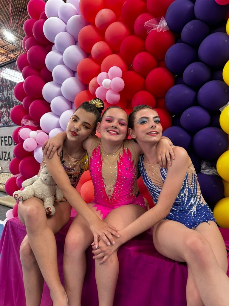 Three young gymnasts in sparkly leotards sit close together on a pink bench in front of a colorful balloon arch, arms around each other; one holds a white teddy bear.