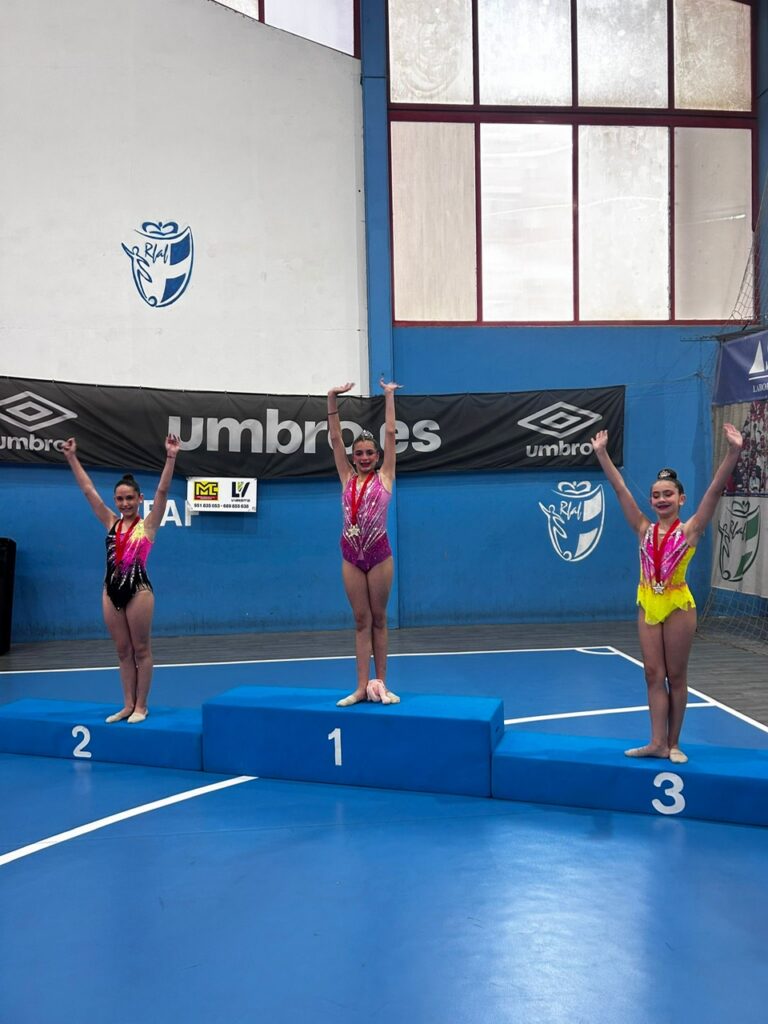Three young gymnasts stand on a blue winners' podium (1st center, 2nd left, 3rd right) with medals, arms raised in celebration inside a sports hall with banners.