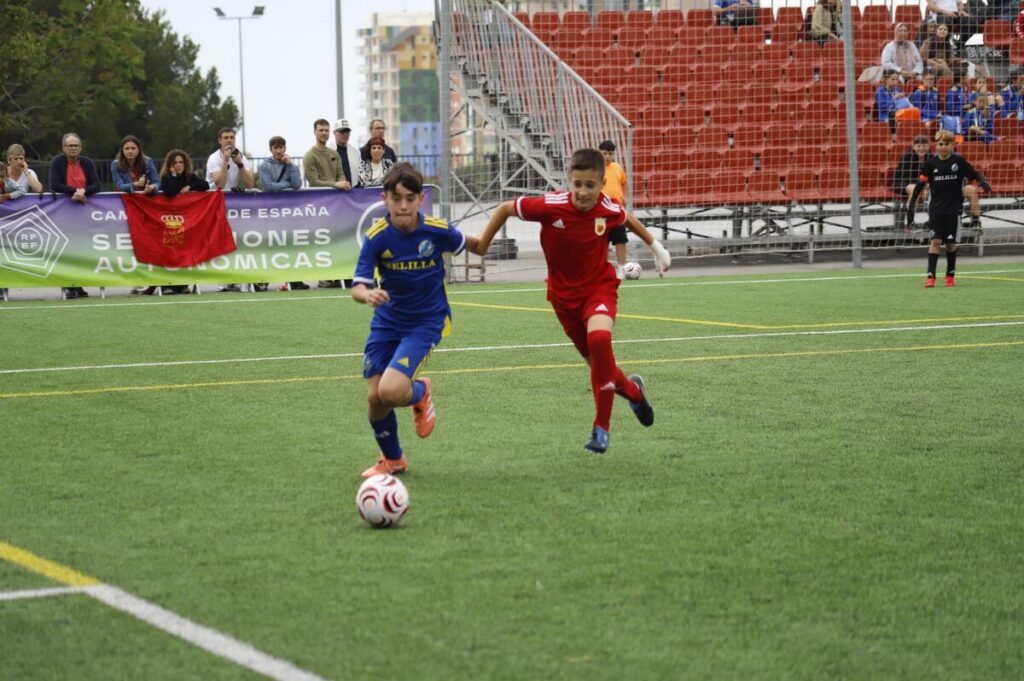 Two youth soccer players compete for the ball on a green artificial turf field as spectators watch from the stands behind a fence and banners.