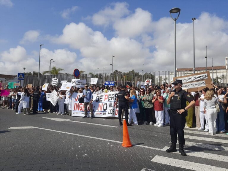Large crowd of protesters on a street holding signs and banners, with a police officer in the foreground and an orange traffic cone nearby.