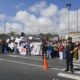 Large crowd of protesters on a street holding signs and banners, with a police officer in the foreground and an orange traffic cone nearby.