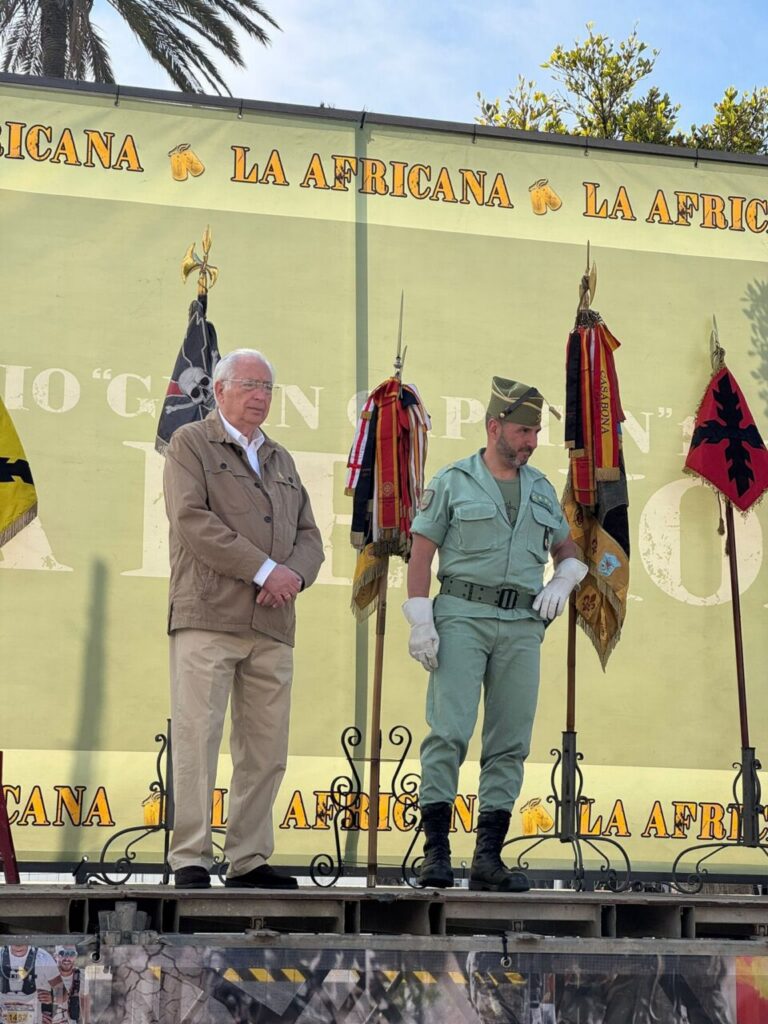 Two men stand on a stage with ceremonial flags; one in a tan jacket and beige pants, the other in a light green military uniform, under a backdrop that reads 'LA AFRICANA'.