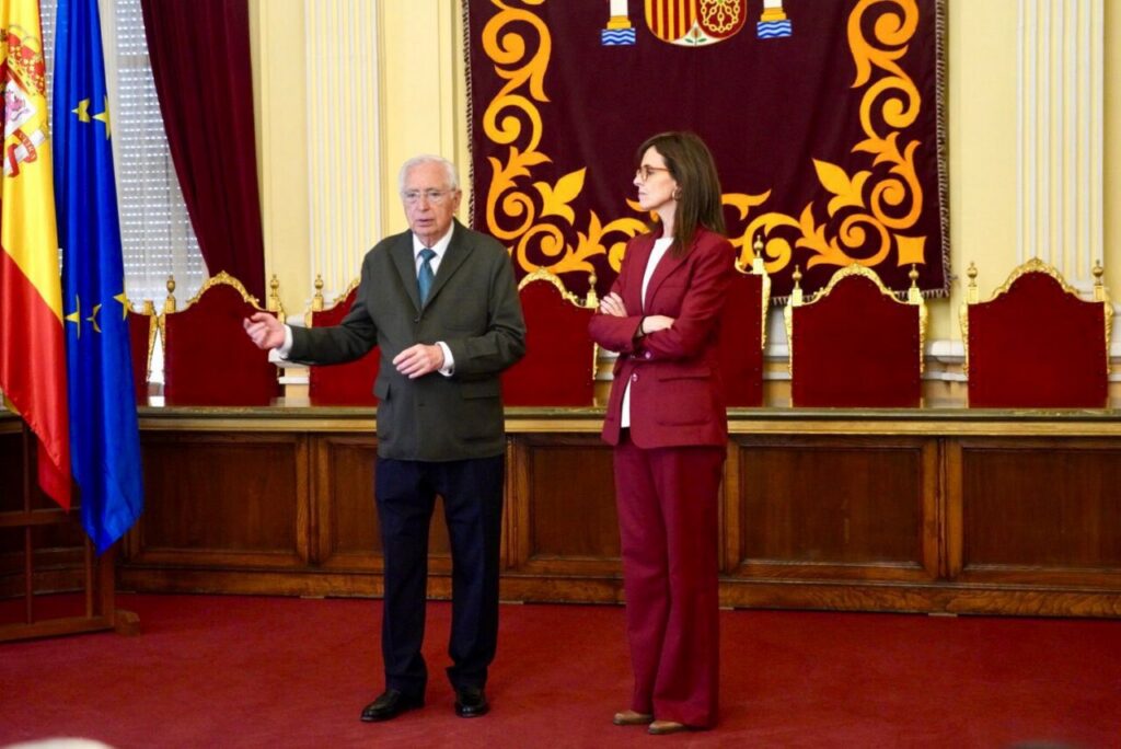 Older man in a dark suit speaks as a woman in a burgundy suit listens in a formal government chamber with flags in the background.