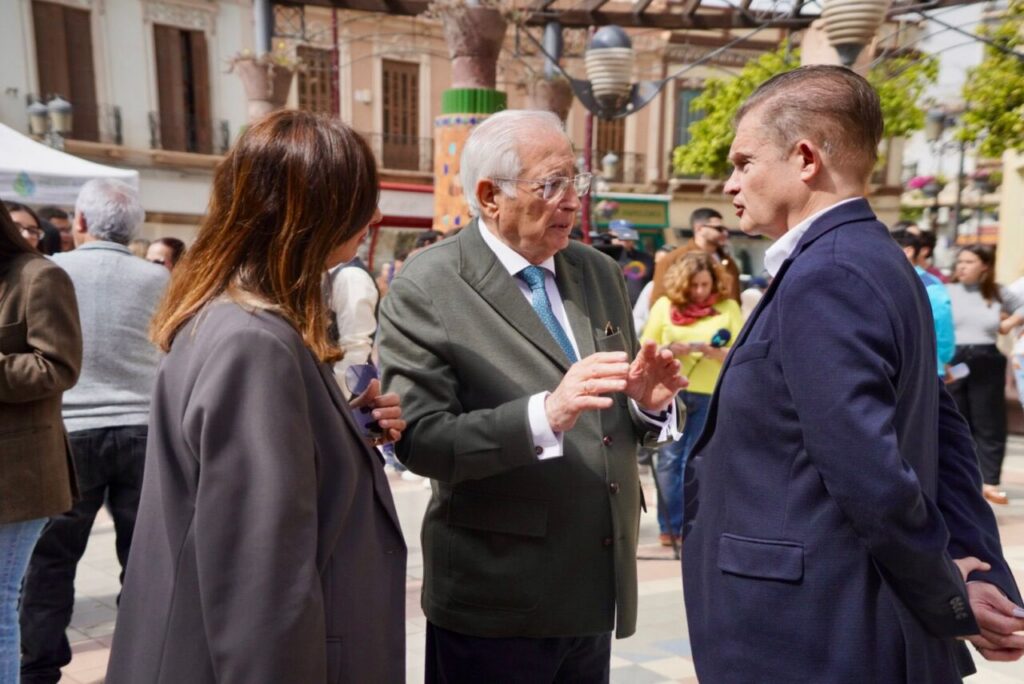 Three adults in a city square conversing: an elderly man in a green blazer speaks to a man in a blue suit, while a woman in a gray coat listens nearby.