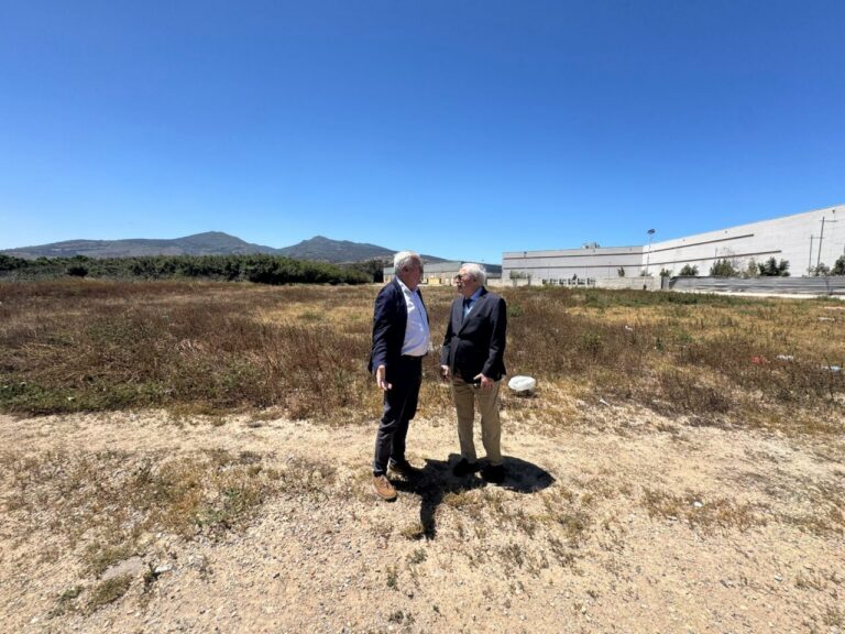 Two men in suits talk in a dry, weed-filled lot beside a large industrial building, with mountains and a blue sky in the background.