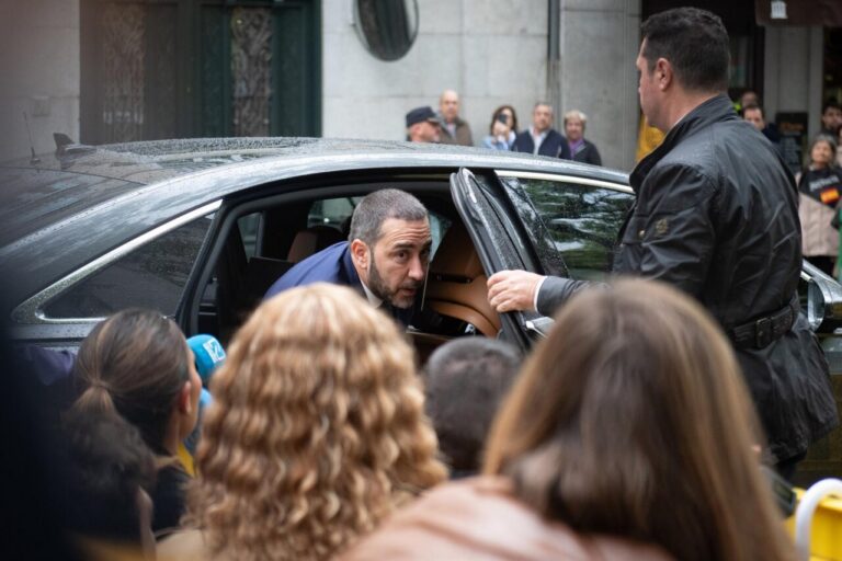 Man leaning into a black car as reporters and onlookers gather around him.
