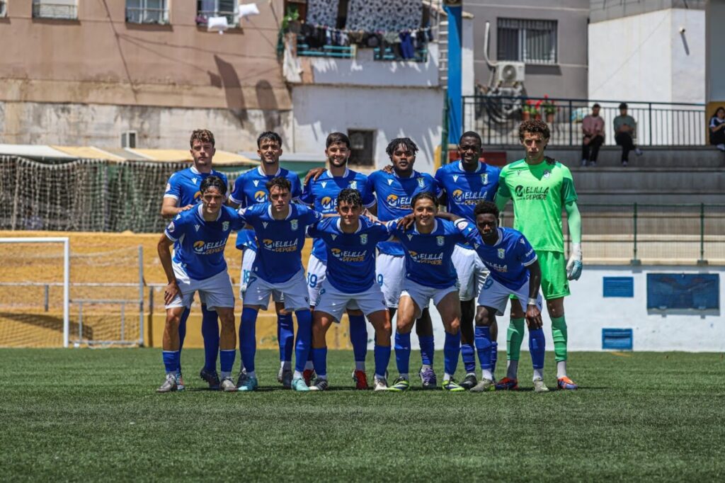 Soccer team in blue uniforms poses for a group photo on a sunny field; goalkeeper in green on the right.