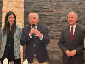 Center: an older man in a navy blazer speaks into a microphone, flanked by a woman in a denim jacket and a man in a dark suit.