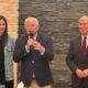 Center: an older man in a navy blazer speaks into a microphone, flanked by a woman in a denim jacket and a man in a dark suit.