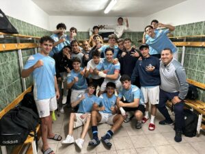 A large group of young soccer players in light-blue jerseys posing together in a green-tiled locker room, some making fists or peace signs.