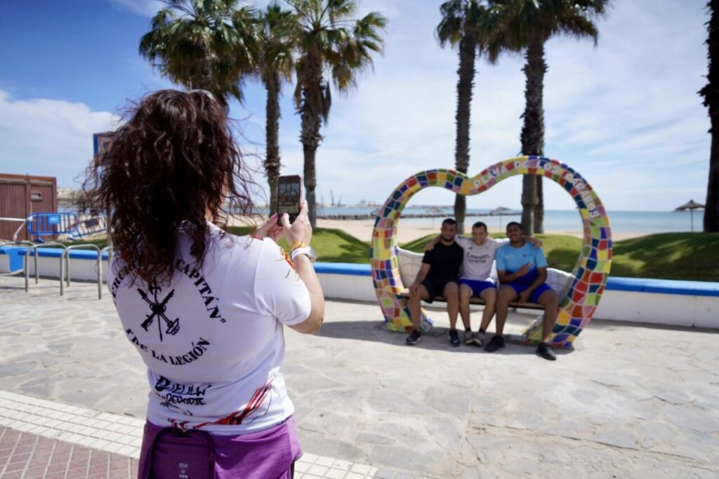 Woman with a phone taking a photo of three men sitting inside a colorful heart-shaped sculpture on a beach promenade. Palm trees and ocean in the background.