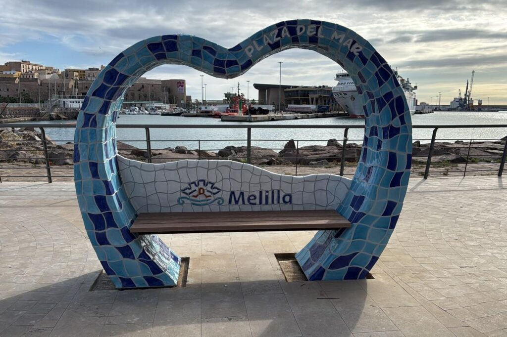 Blue mosaic heart-shaped bench by Melilla's waterfront, with a wooden seat and harbor in the background.