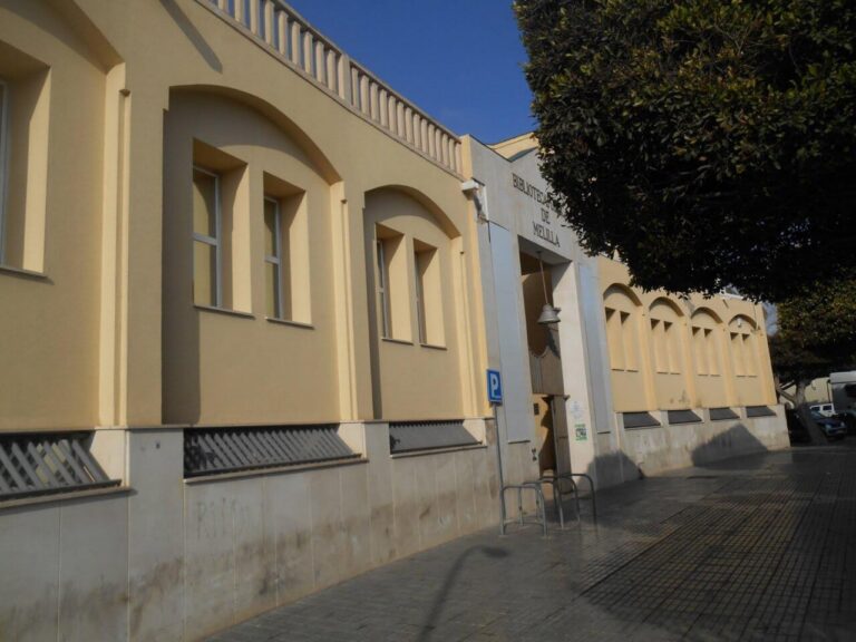 Exterior view of the Biblioteca de Melilla, a pale yellow building with arched windows along a sidewalk under a blue sky.