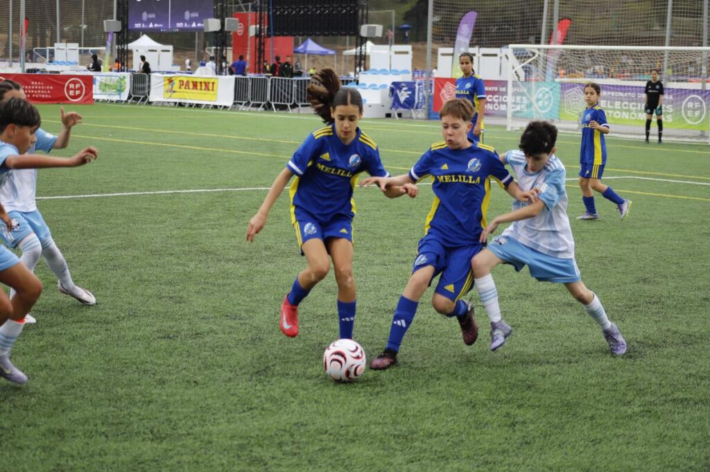 Girls' youth soccer match: two players in blue uniforms contest the ball as others chase nearby on a sunny artificial turf field with a goal and banners in the background
