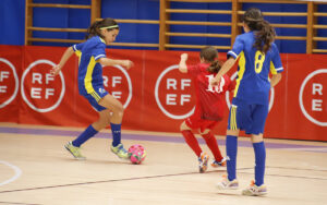 Three young players compete in an indoor futsal match: two in blue jerseys defend while a red-jersey player advances toward the ball on a gym floor.