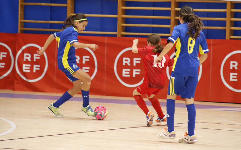 Three young players compete in an indoor futsal match: two in blue jerseys defend while a red-jersey player advances toward the ball on a gym floor.