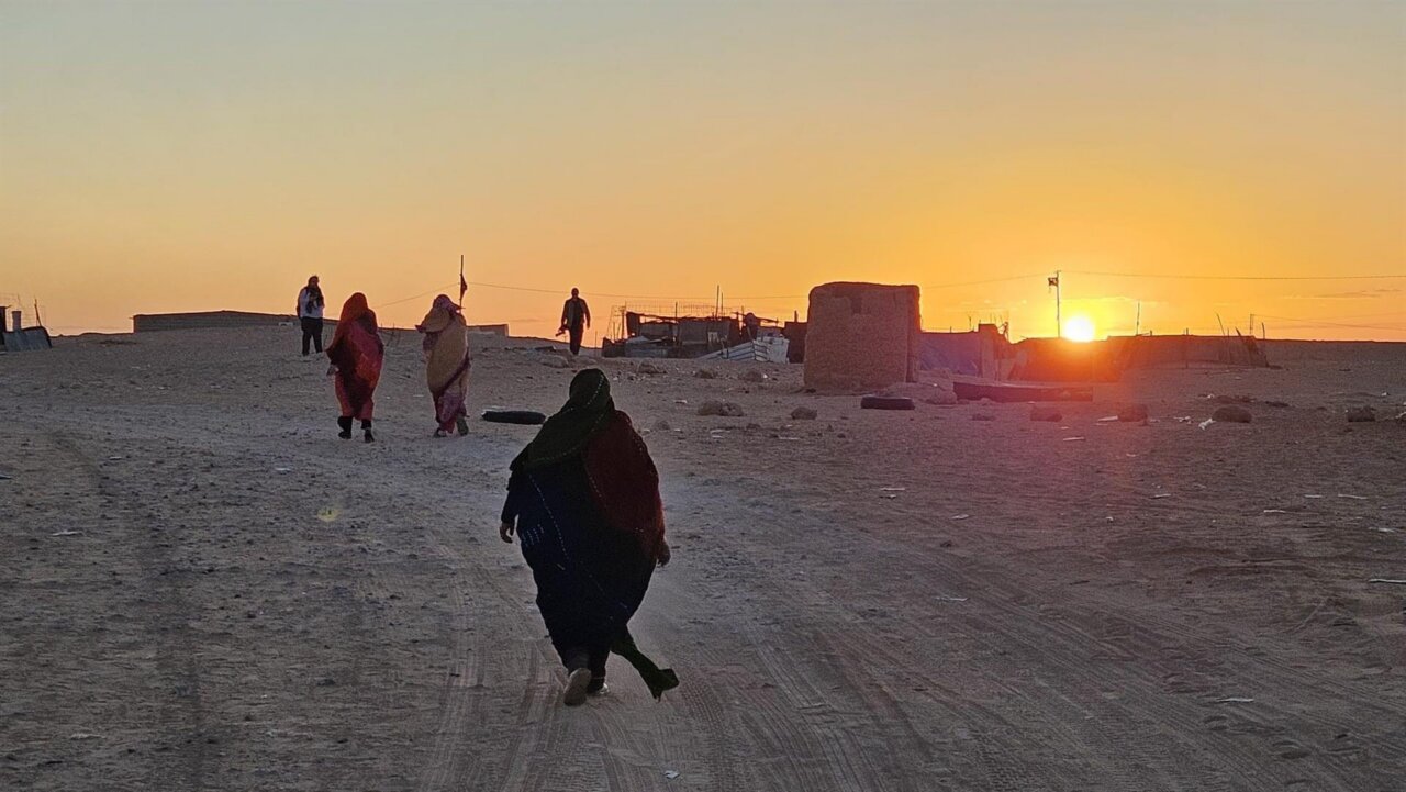 People in traditional clothing walk along a dusty desert road toward a small settlement at sunset, orange sky.