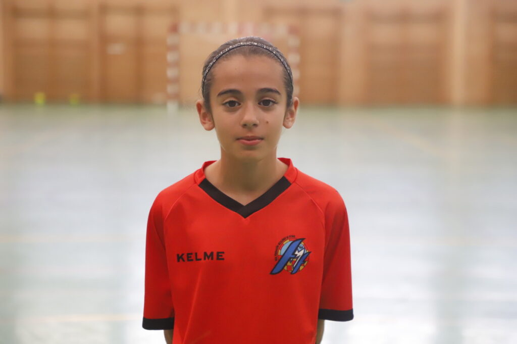 Young girl in a red sports jersey standing in a gym, facing the camera with a neutral expression.