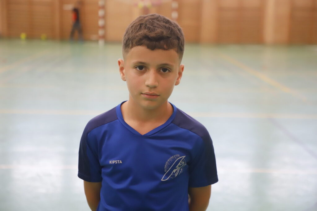 Young boy in a blue sports jersey stands in an indoor gymnasium, looking at the camera.