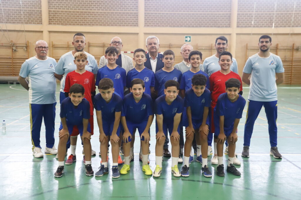 Group of young athletes in blue and red uniforms posing for a team photo in a gym, with coaches standing behind them.