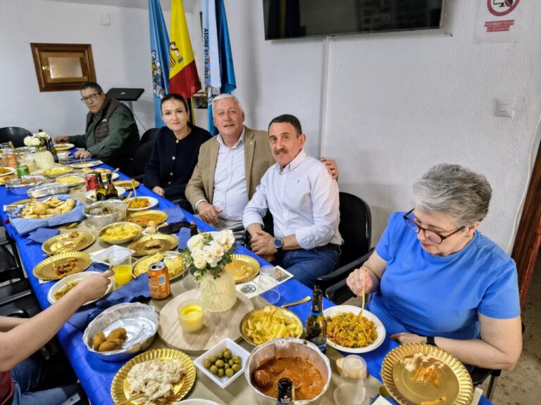 Five adults sit along a long table covered with a blue tablecloth, sharing a meal with plates, bowls, bread, and drinks in a festive setting with flags in the background and a TV on the wall behind them.