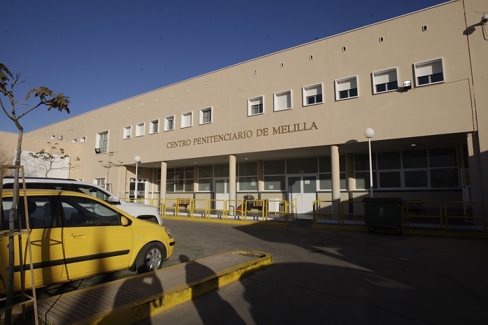 Exterior of Centro Penitenciario de Melilla, a beige prison building with its name on the façade; a yellow taxi is parked outside.