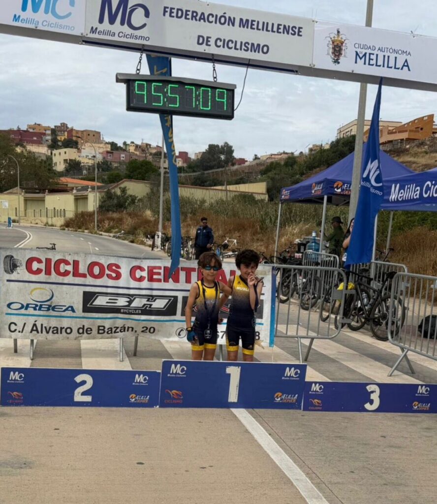 Two young cyclists stand on the winners' podium at the finish line of a cycling race, wearing matching yellow and black outfits with banners in the background.