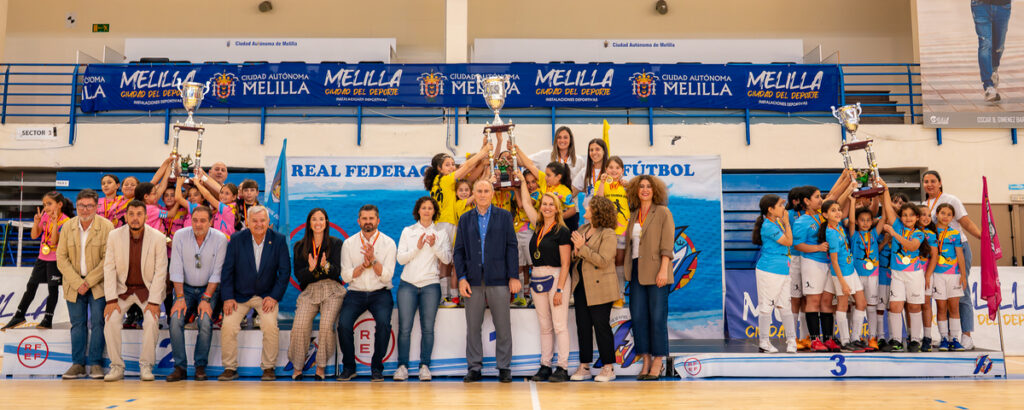 Group of young athletes and officials celebrate on a podium with trophies at a futsal event, Real Federación banner behind them.
