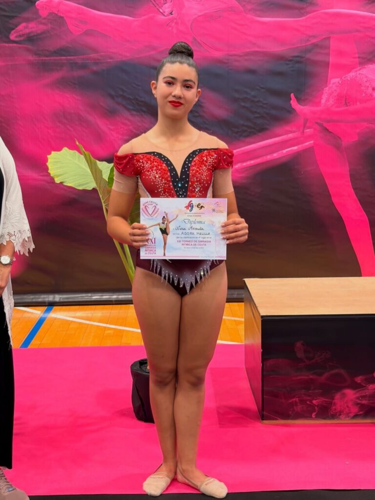 Young female gymnast in a red and black sparkly leotard stands on a pink podium, holding a diploma certificate.